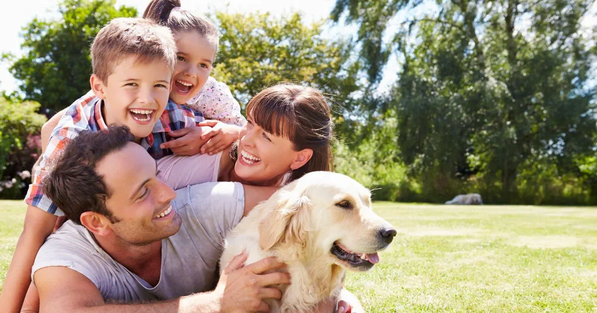 a family, mom, dad and two young children with a labrador dog all in an embrace oustide in the park