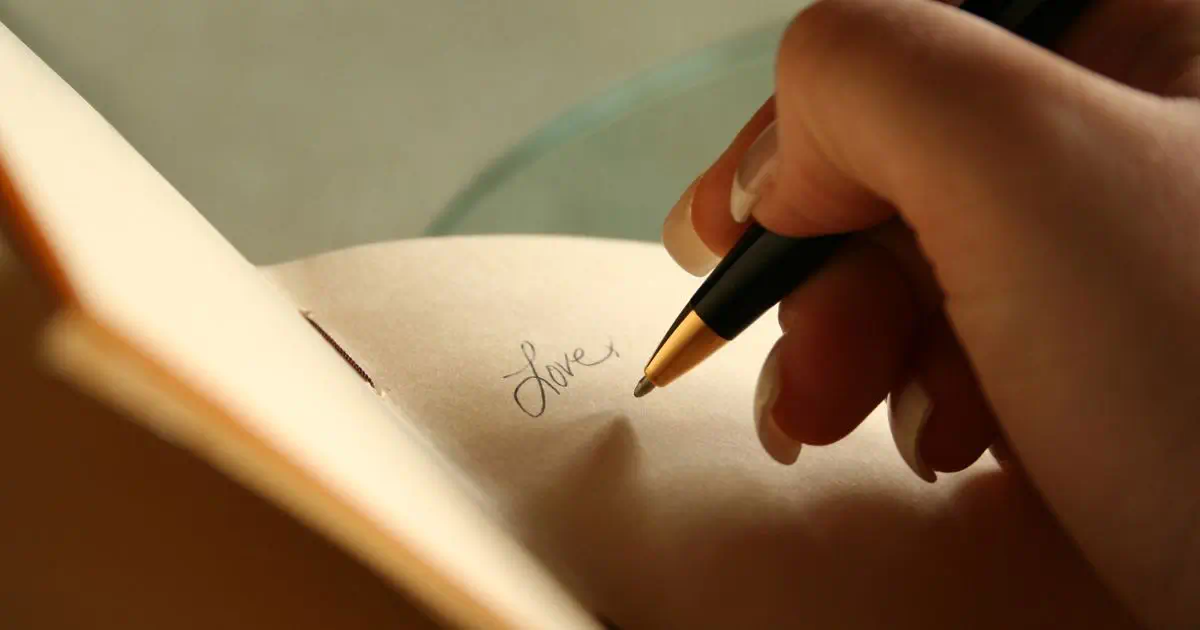 an open journal with the word love written on it. A hand holding a pen hovering over the page